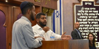 Photo of two men with short dark hair and goatees, wearing yarmulkes. One man is speaking behind a podium in a synagogue. There is an Israeli flag and Hebrew text in the background.