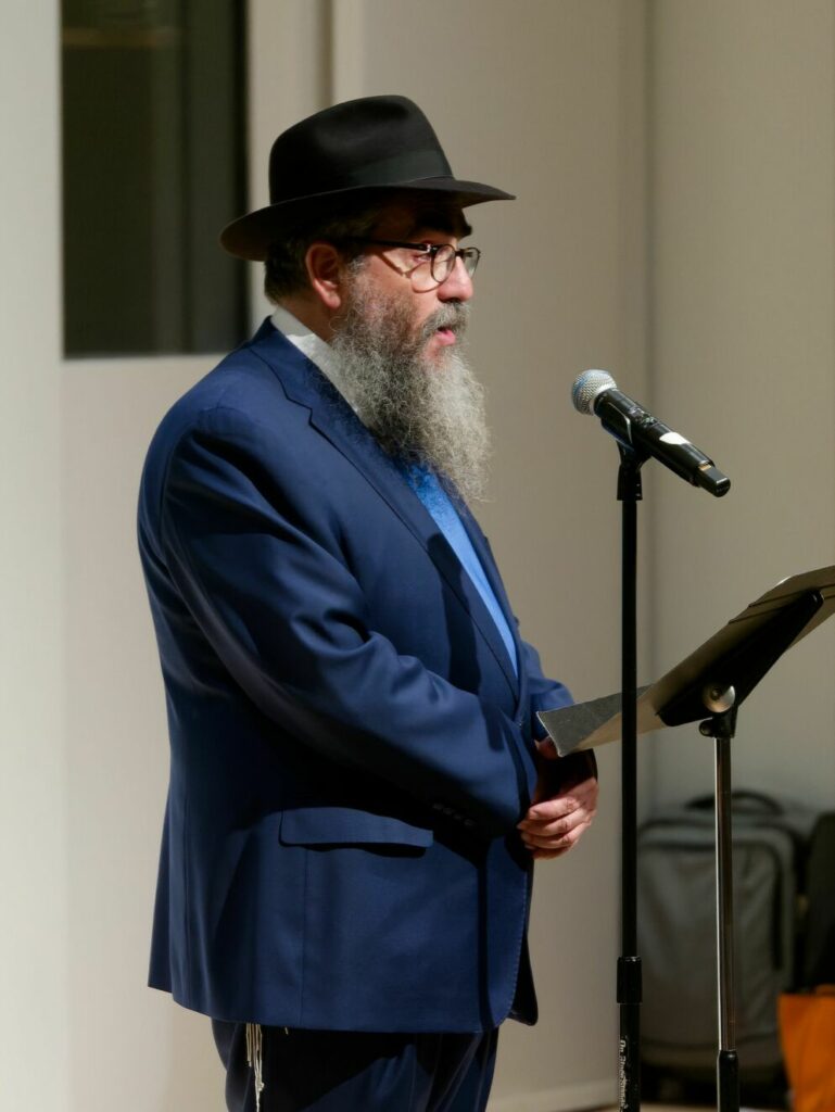 Photo of a bearded man standing at a microphone stand. He wears a black hat, suit and wire-rimmed classes.