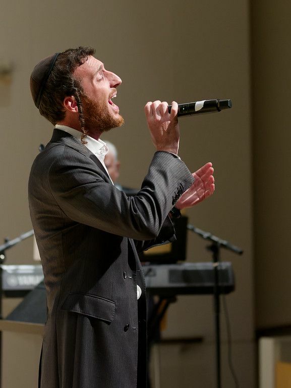 Side profile of an Orthodox Jewish man singing into a microphone that he holds in one hand. His eyes are closed and his mouth is open mid-song.