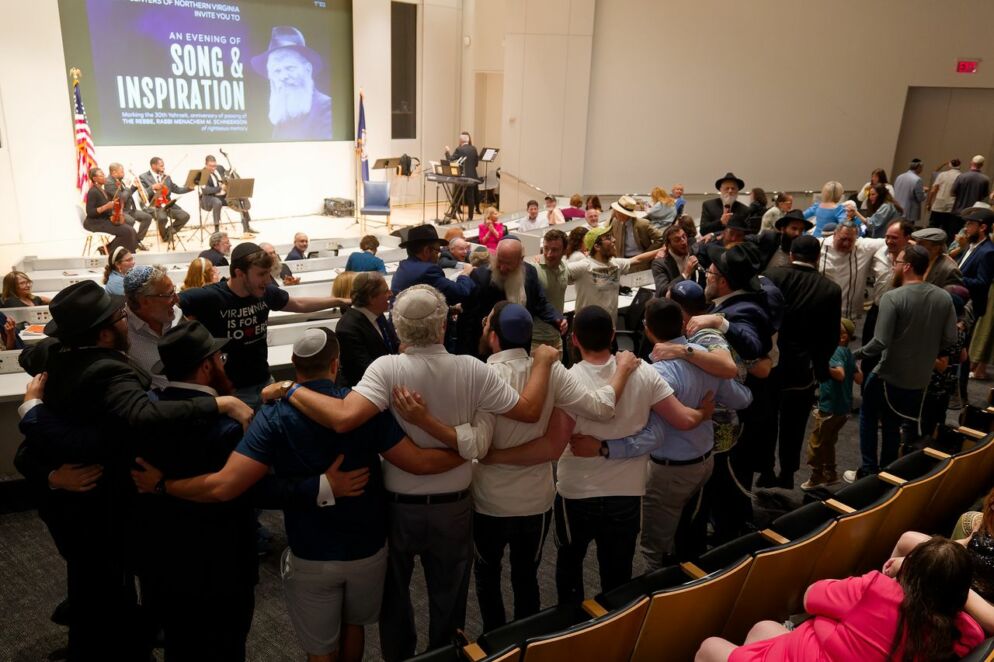 Dozens of people with their arms around each other form a circle in an auditorium. In front of the people is a screen that reads "An Evening of Song & Inspiration."