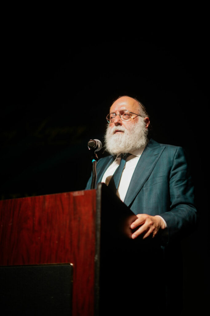 A man with wire-rimmed glasses and a white beard stands behind a podium.
