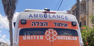 Photo of a teen girl with short dark brown hair smiling in front of the back of an Israeli ambulance. She is wearing khaki pants, a black T-shirt and a neon orange vest.