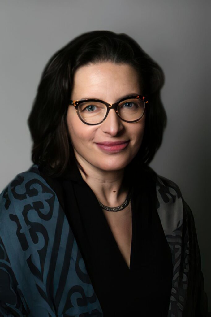Headshot of a woman with shoulder-length straight brown hair and dark-colored glasses. She is wearing a black and gray patterned V-neck top and a necklace.