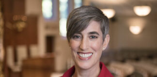 Headshot of a woman smiling at the camera inside a synagogue. Her graying short hair is styled in a pixie cut and she is wearing a red top with a cowled collar. She is smiling at the camera. The background is blurred.