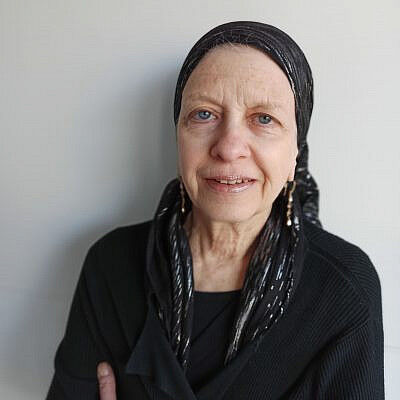 Headshot of a woman wearing black with a black scarf around her head. She is smiling at the camera in front of a plain gray background.