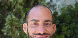 Photo of a man with a buzz cut and a dark goatee smiling in front of greenery. He is wearing a navy blue blazer over a white shirt checkered with light pink and a light blue tie.