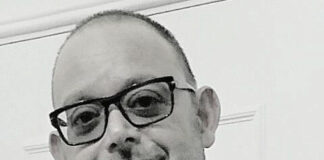 Headshot of a man with a buzz cut and black glasses wearing a black shirt. He is smiling at the camera.