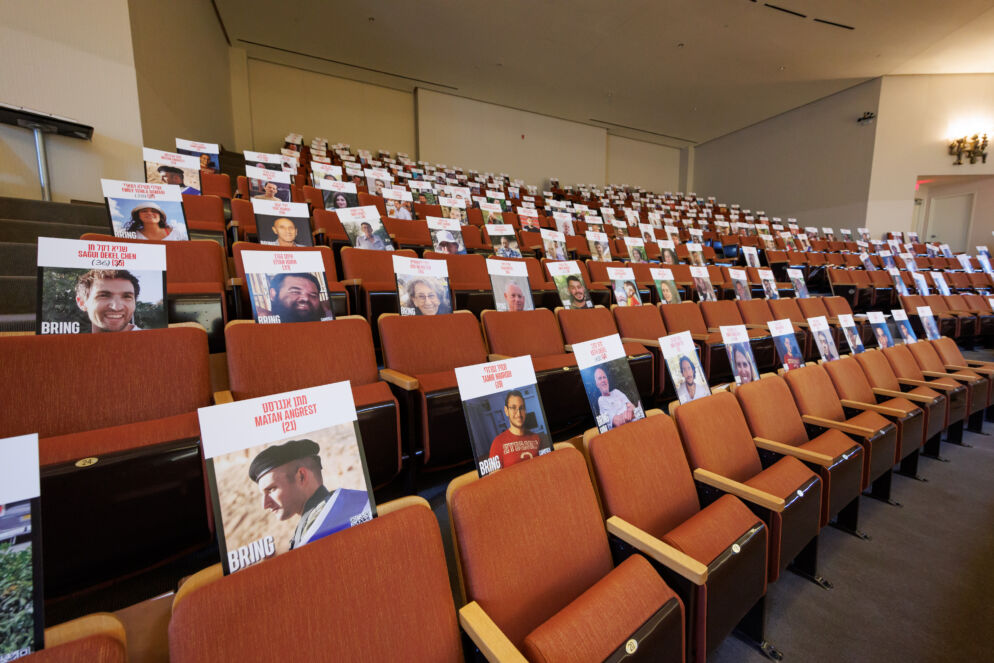 Photo of rows of empty reddish-brown theater-style chairs. On each chair is a poster containing the name, age and photo of a hostage.