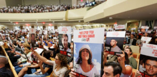 Photo of hundreds of people solemnly sitting indoors and holding up signs with hostages' names and photos. The room is packed.