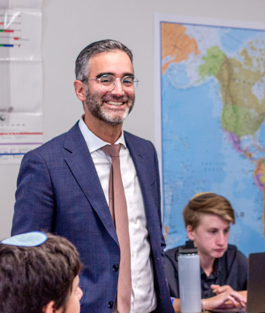 Photo of a man with gray hair, glasses and a gray goatee smiling and standing in a day school classroom. There is a world map hanging on the wall behind him.
