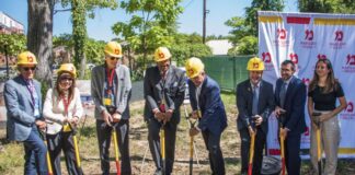 Photo of eight people wearing yellow construction hats and posing with red shovels containing the white Hillel logo outdoors.