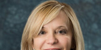 Headshot of a woman with straight blonde hair a little past her shoulders. She is wearing a white beaded necklace, a black top and is smiling at the camera.