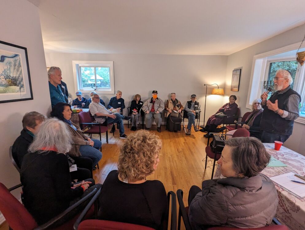 Photo of a rabbi with white hair speaking into a microphone to about two dozen adults sitting in a large circle indoors .