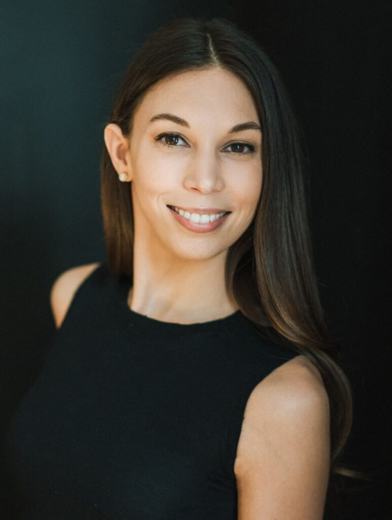 Headshot of a young woman with long straight brown hair. She is wearing a sleeveless black top and smiling at the camera.