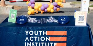 Photo of a navy blue tablecloth draped over a table that reads "Youth Action Institute" in white text. Below that in smaller text reads "Grow leadership skills," "volunteer with purpose," "earn service learning hours" and "make new friends." There is a balloon wreath of yellow and purple balloons behind the table.
