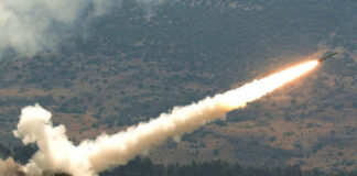 Photo of a rocket firing at a grassy ground. There are large white clouds around the rocket.