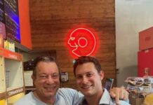 Bethesda Bagels Wins Best Bagels 2024 Photo of two men smiling at the camera from inside a bagel shop. The older man has his arm around the younger man, who is holding a sesame bagel. They are both wearing gray athletic T-shirts.