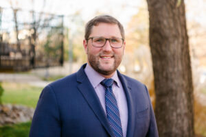 Headshot of a man with glasses smiling at the camera outside. He is wearing a navy blue suit with a light purple shirt and dark blue tie.
