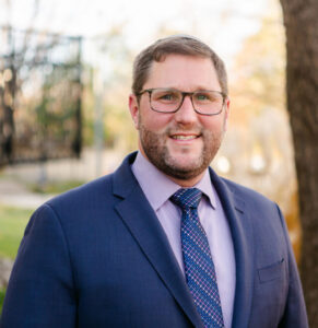Headshot of a man with glasses smiling at the camera outside. He is wearing a navy blue suit with a light purple shirt and dark blue tie.