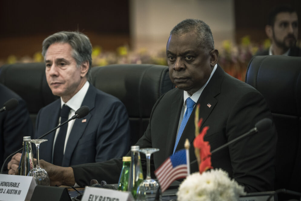 Photo of two men sitting side by side at a table looking serious. They are both wearing suits.