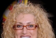 Hazzan Kim Komrad Shatters Glass Ceilings Headshot of a woman with long wavy blonde hair and glasses. She is smiling at the camera.