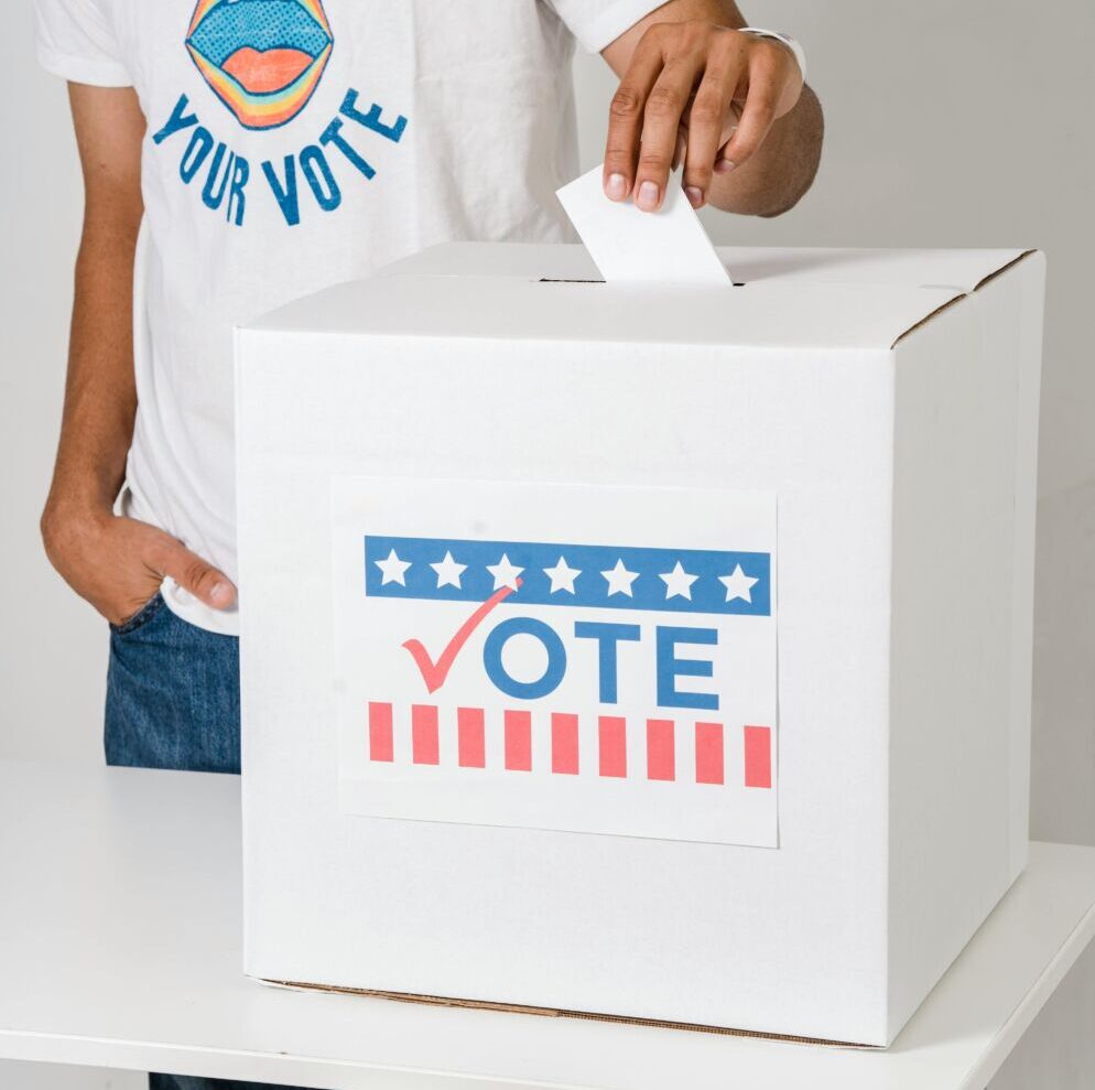 Stock photo of a man putting a folded white piece of paper into a ballot box that says "Vote" in red, white and blue on its front. The man is wearing a white T-shirt that reads "Your Voice, Your Vote" in blue text in a circle around an illustration of an open mouth.