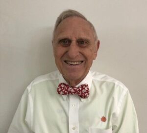 Headshot of an older man smiling at the camera against a white background. He is wearing a white button-down shirt and a red patterned bow tie.