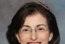 Headshot of a woman with short dark hair cut into a bob and glasses smiling at the camera. She is wearing red lipstick and a headband on her head.