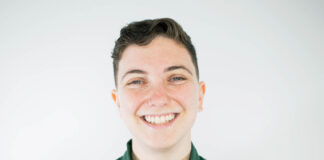 Photo of a person with short brown hair smiling at the camera against a white background. They are wearing a green button-down shirt.