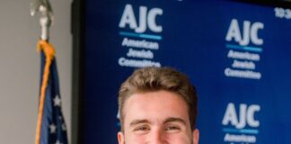 Headshot of a young man with short brown hair and some facial hair smiling with his arms crossed in front of him. He is wearing a suit with a purple and blue tie and he is standing in front of an American flag and the American Jewish Committee blue backdrop.