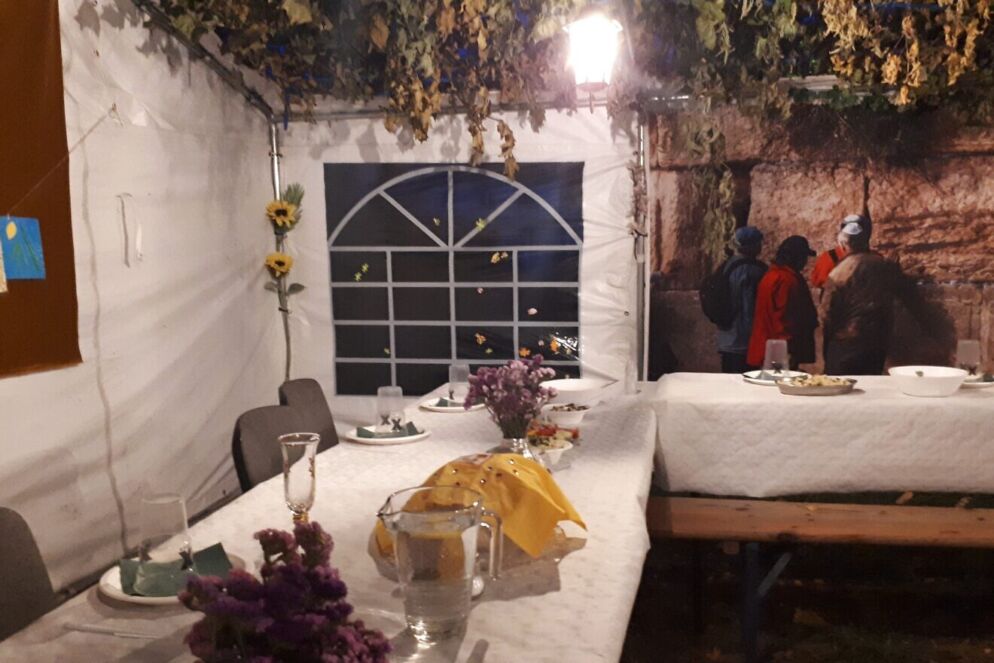 Photo of the inside of an elaborately decorated sukkah. There is a dining area in front of a window and lots of flowers hanging from the ceiling.