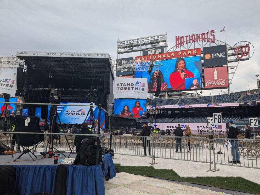Photo from the center of the field at Nationals Park. An Indigenous woman with long straight dark hair and dangling beaded earrings is shown on a large screen and smaller screen underneath the Nationals sign.