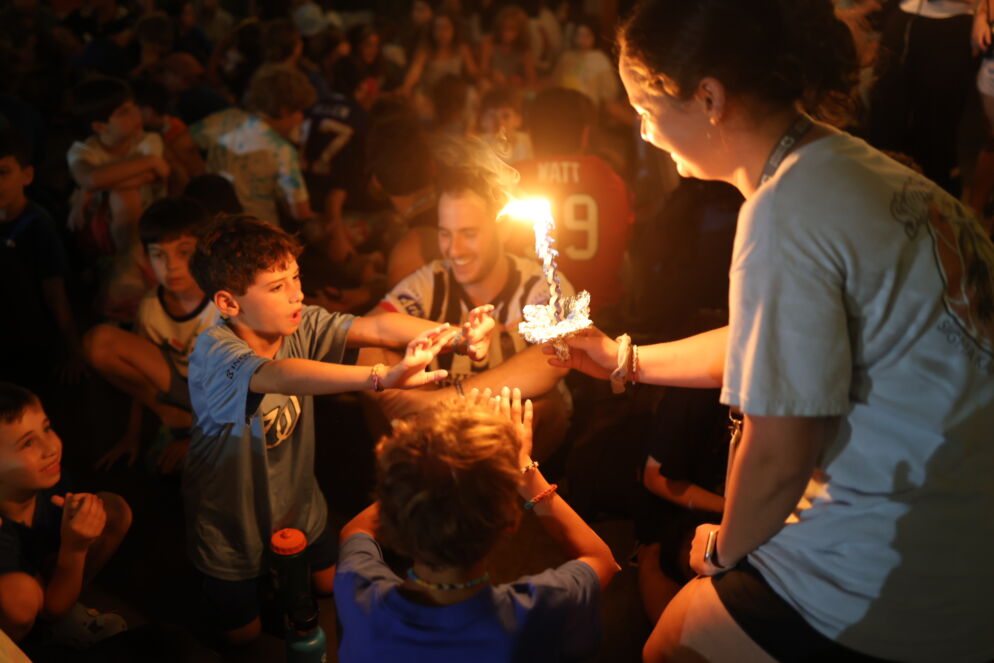 Photo of kids gathered around a lit braided candle that a teen camp counselor is holding in the dark.