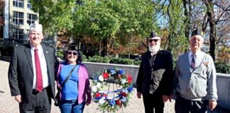 Photo of four adults; two standing outside on a stone walkway on either side of a wreath with red, white and blue flowers. The man on the far left is dressed in a black suit and red tie, the woman next to him is wearing a blue shirt, purple sweater and jeans, and the two men on the right are in military uniform.
