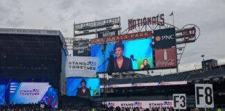 Photo from the center of the field at Nationals Stadium. Dozens of rows of folding chairs are set up. A Black woman with her braided locks pulled back is projected on two screens. She is speaking into a microphone from the stage.