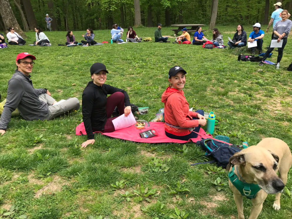 Photo of three adults sitting on a pink picnic blanket on the grass. They are wearing baseball caps and long-sleeved athletic wear. There is a tan medium-large dog in the lower left corner.