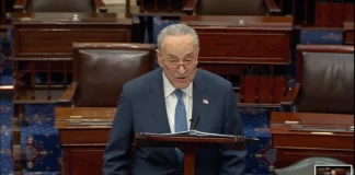 Photo of a man with short gray hair and glasses standing behind a podium speaking. He is wearing a suit with a blue tie.