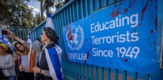 Photo of people with the Israeli flag draped around their shoulders protesting in front of a large blue sign with the UNRWA logo in white and the text "Educating Terrorists Since 1949."