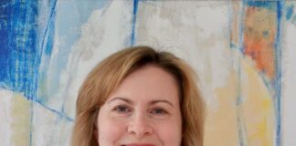 Headshot of a woman with short light brown hair wearing a blue top and a necklace. She is smiling at the camera in front of a blue and yellow painting.