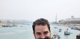 Photo of a 37-year-old man with dark brown hair and a brown goatee smiling at a camera in front of a body of water with about half a dozen boats. He is wearing a navy blue suit with a tie.