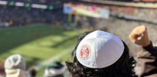 Photo of the back of someone's head wearing a white yarmulke with the Commanders' maroon logo on it. The person has short curly dark hair and is wearing a maroon hoodie in the stands of a football stadium, watching the game.