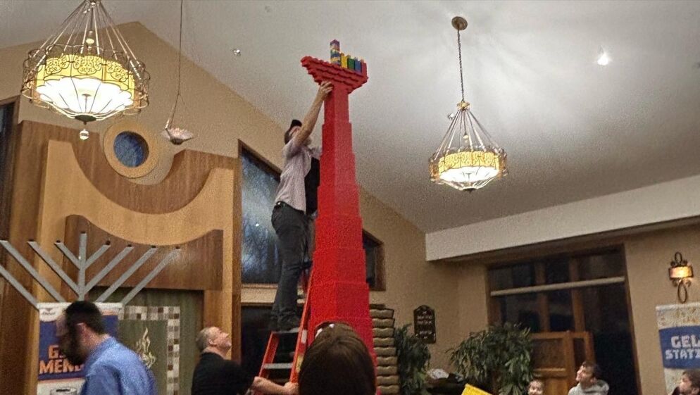 Photo of a person standing on a ladder to place candles atop a tall red LEGO brick structure in the shape of a menorah inside a building. There is a lit chandelier hanging next to the structure.