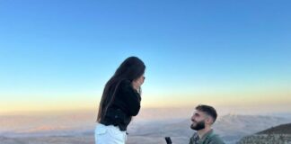 Photo of a man in an army uniform kneeling on one knee in front of a young woman with long, dark hair who has both hands covering her mouth in surprise.