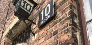 Photo of a blonde woman standing outside a brick building with one hand resting on a concrete windowsill. She is wearing a black coat and a navy blue patterned scarf.