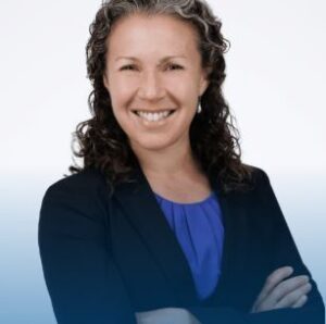Headshot of a middle-aged woman with curly brown hair smiling at the camera. She is wearing a blue shirt under a black blazer and crossing her arms in front of her.