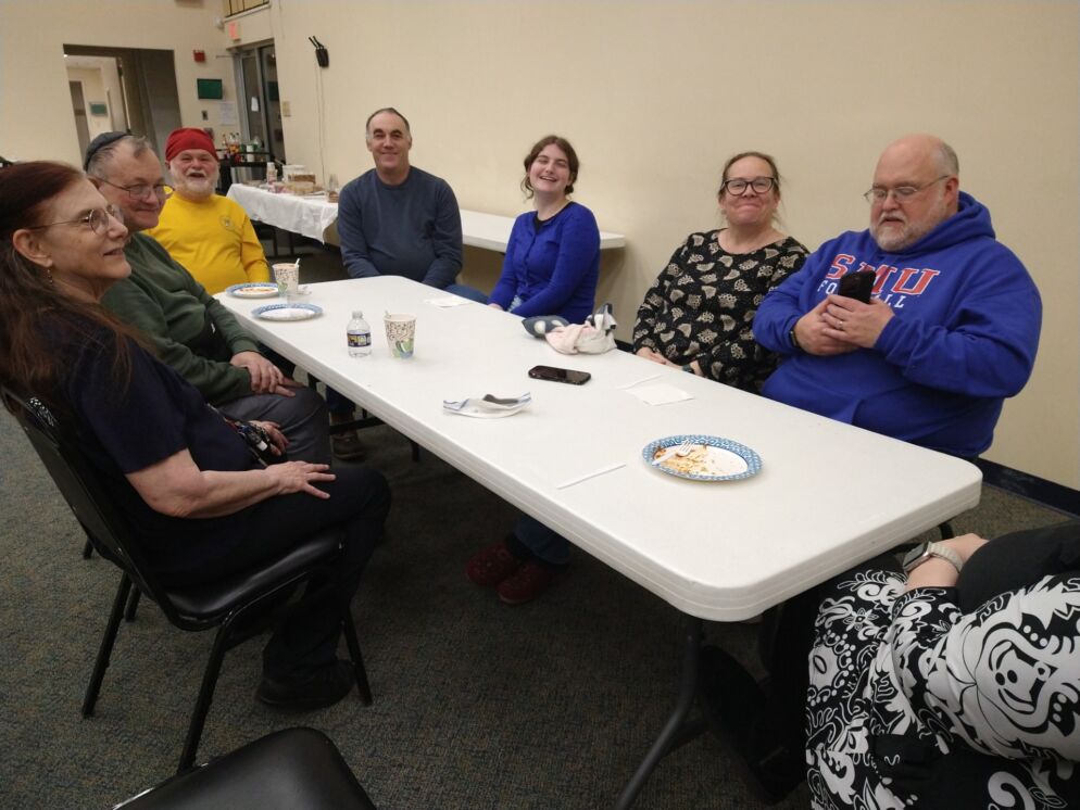 Photo of half a dozen people seated around a rectangular white folding table inside a room.