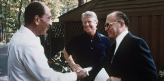 Photo of two men in suits shaking hands as an older man with short gray hair looks on in the background.