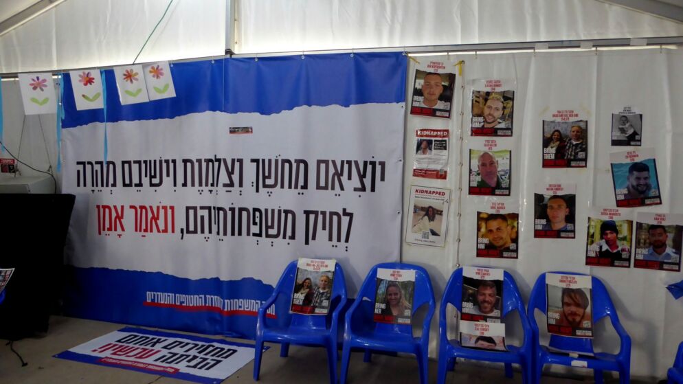Photo of a blue and white banner calling for the release of the hostages in black Hebrew text. There are hostage posters with people's faces on each one.