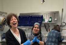 Photo of two women in an industrial kitchen smiling and laughing as they cook. They are dressed in black and wearing blue gloves with a large tub of food on the stainless steel counter in front of them.