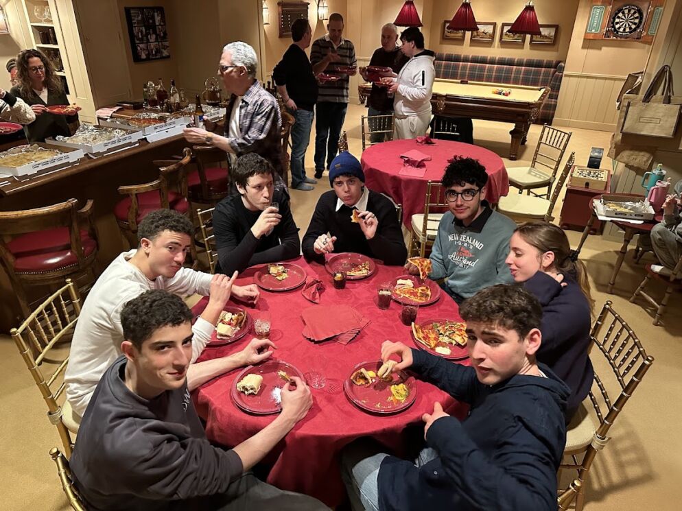 Photo of seven teens sitting around a round table with a red tablecloth. They are eating pizza.